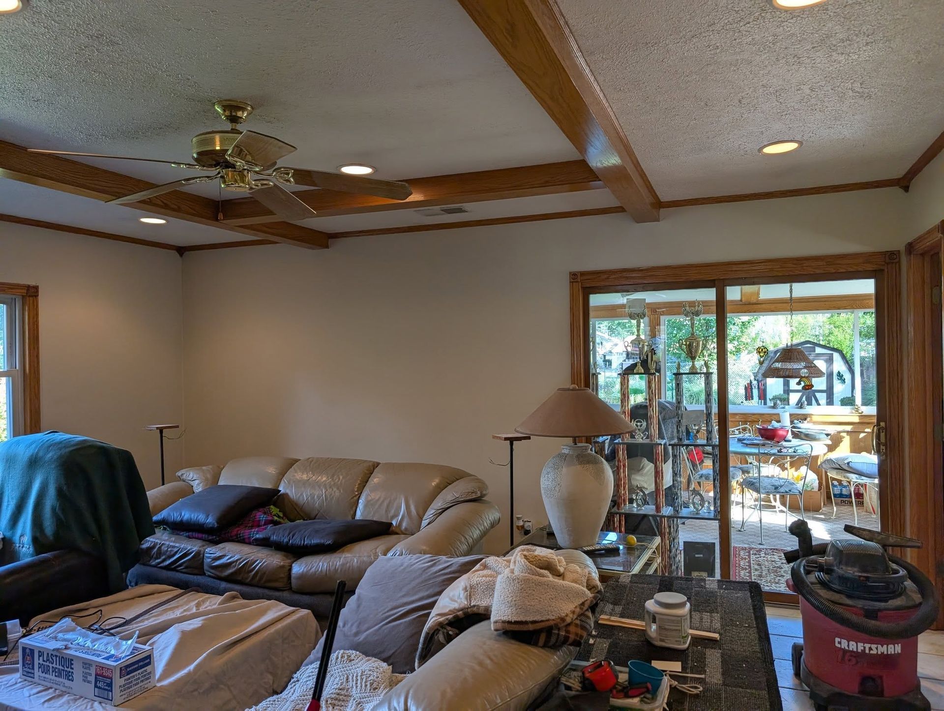 A living room with a beige sofa, wooden ceiling beams, a ceiling fan, and a view through sliding glass doors to a patio.