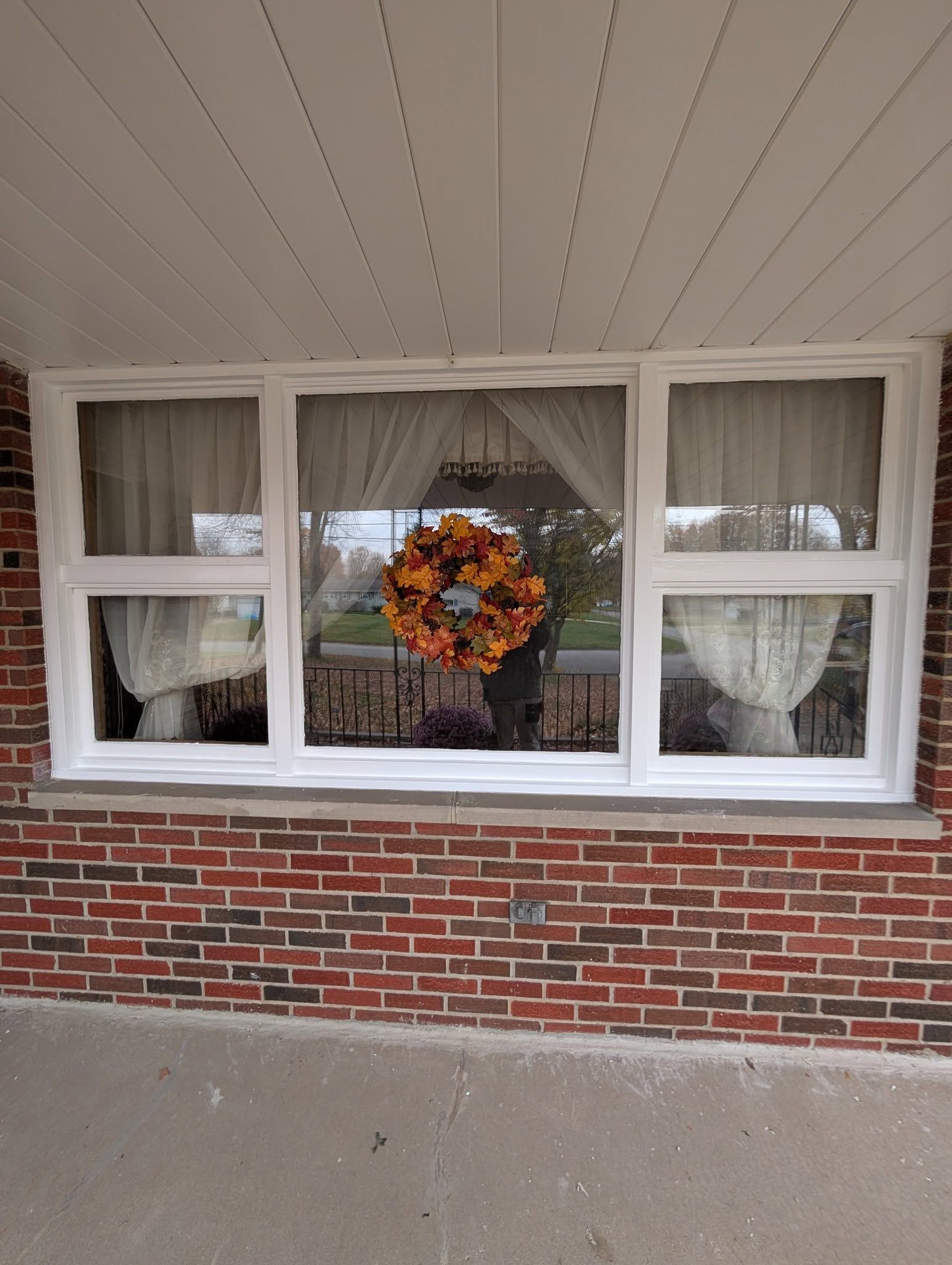 A three-pane window with white frames and lace curtains on a brick exterior wall, featuring a decorative autumn wreath.