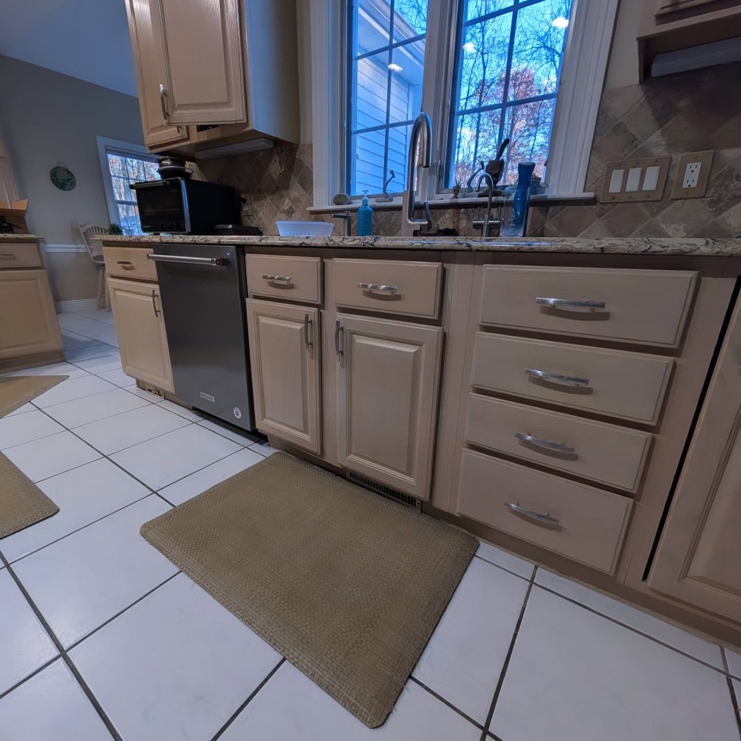 A beige-toned kitchen with a stainless steel dishwasher, granite countertops, and light-colored cabinetry over tile floors.