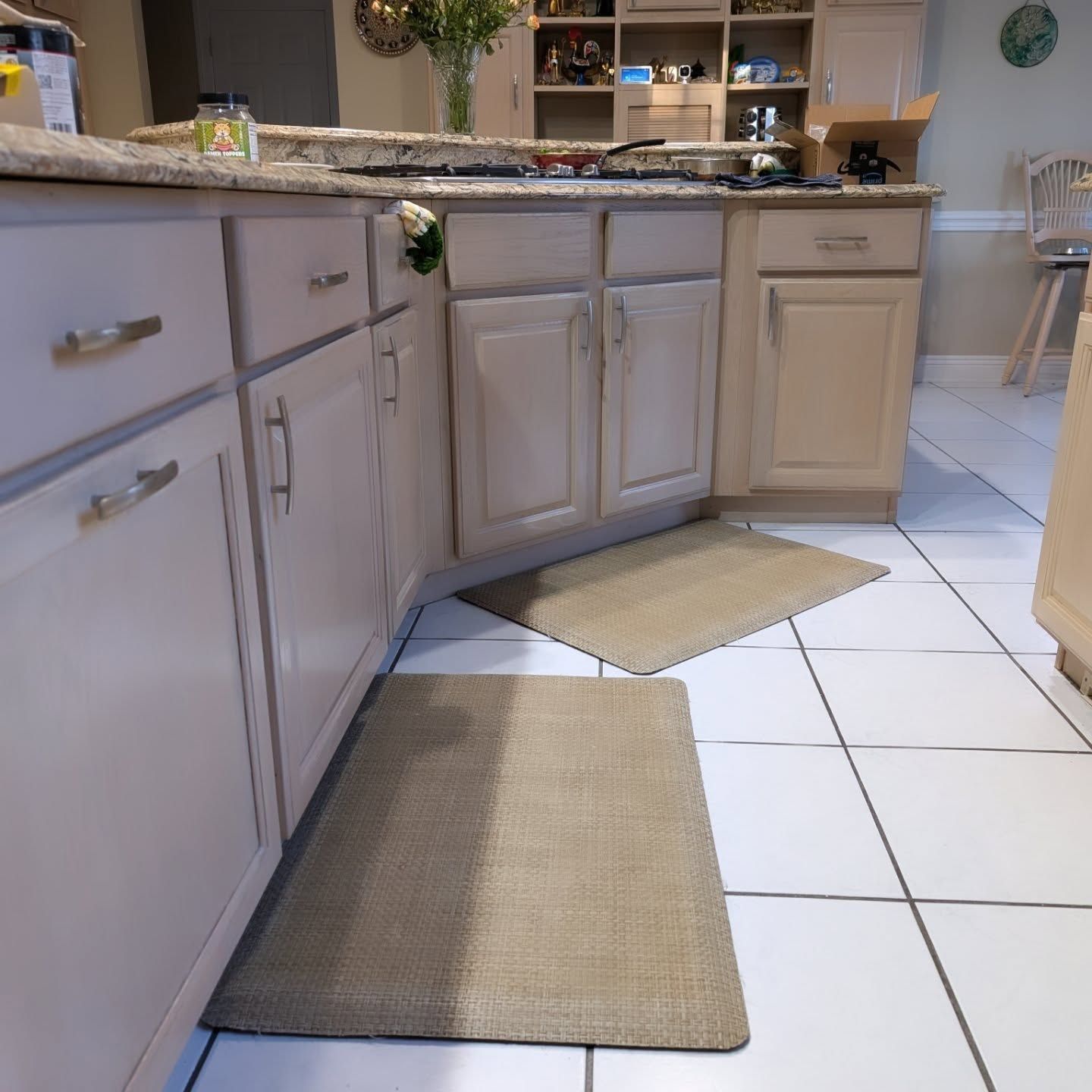 Two beige anti-fatigue mats on a white tiled kitchen floor in front of light-colored kitchen cabinets.