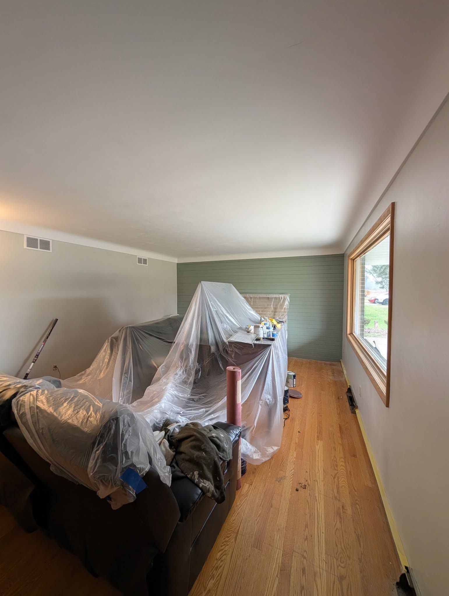 A room under renovation with hardwood floors, covered furniture, and an accent wall being painted green.