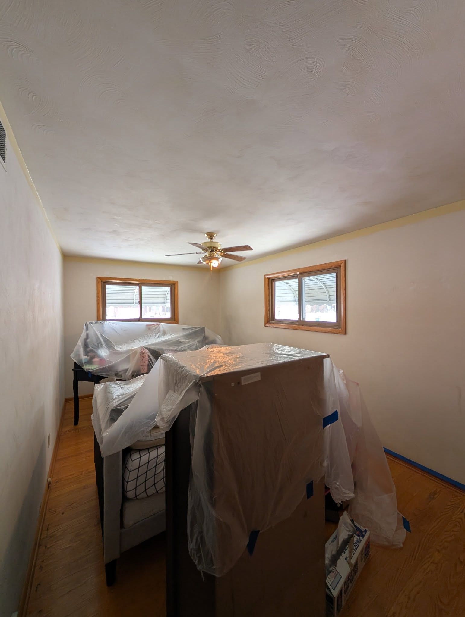 A room with hardwood floors, two windows, and furniture covered with plastic for protection during home renovations.