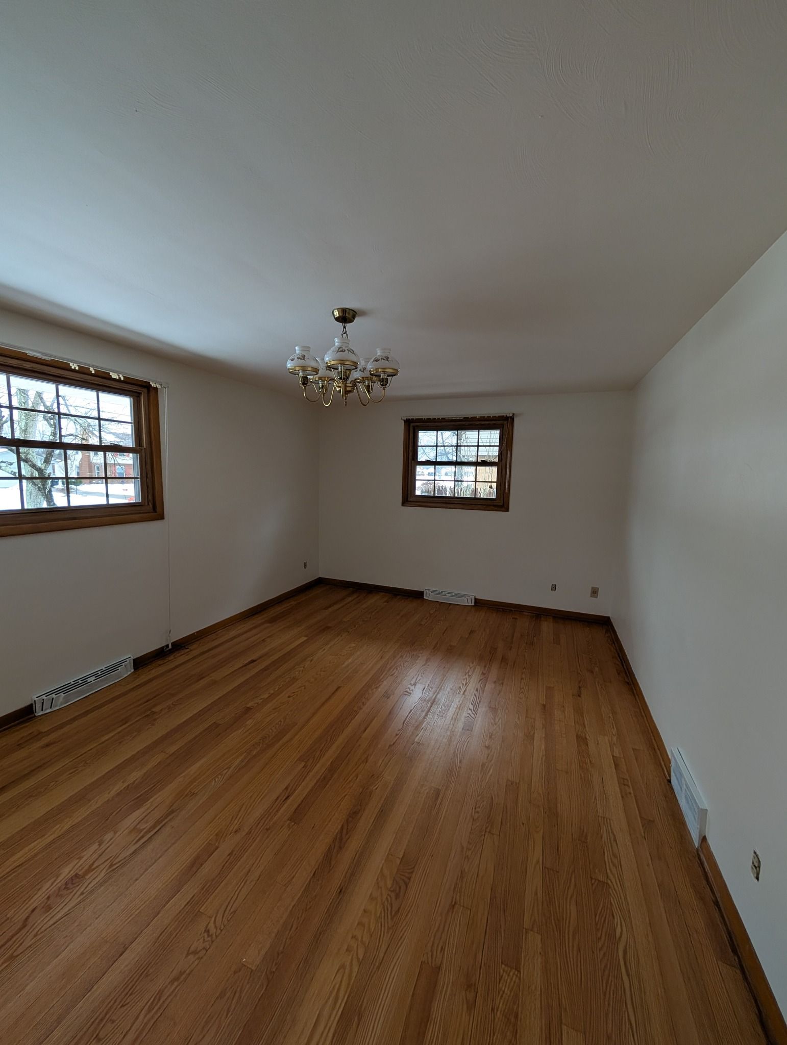 An empty, rectangular room with polished hardwood floors, cream-colored walls, and two windows with wood-toned frames.