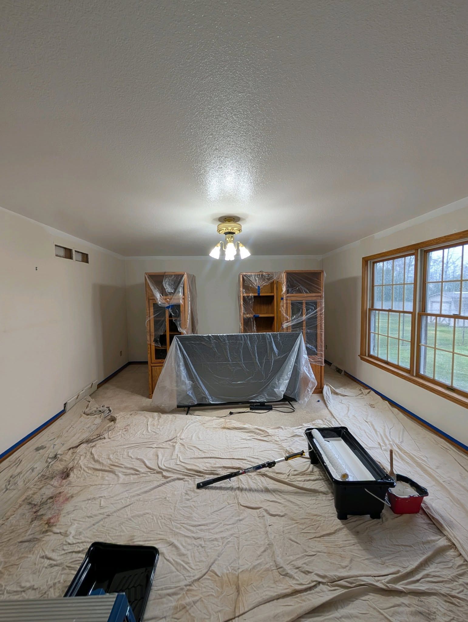 A room under renovation with covered furniture, drop cloths on the floor, and painting supplies, including a roller tray.
