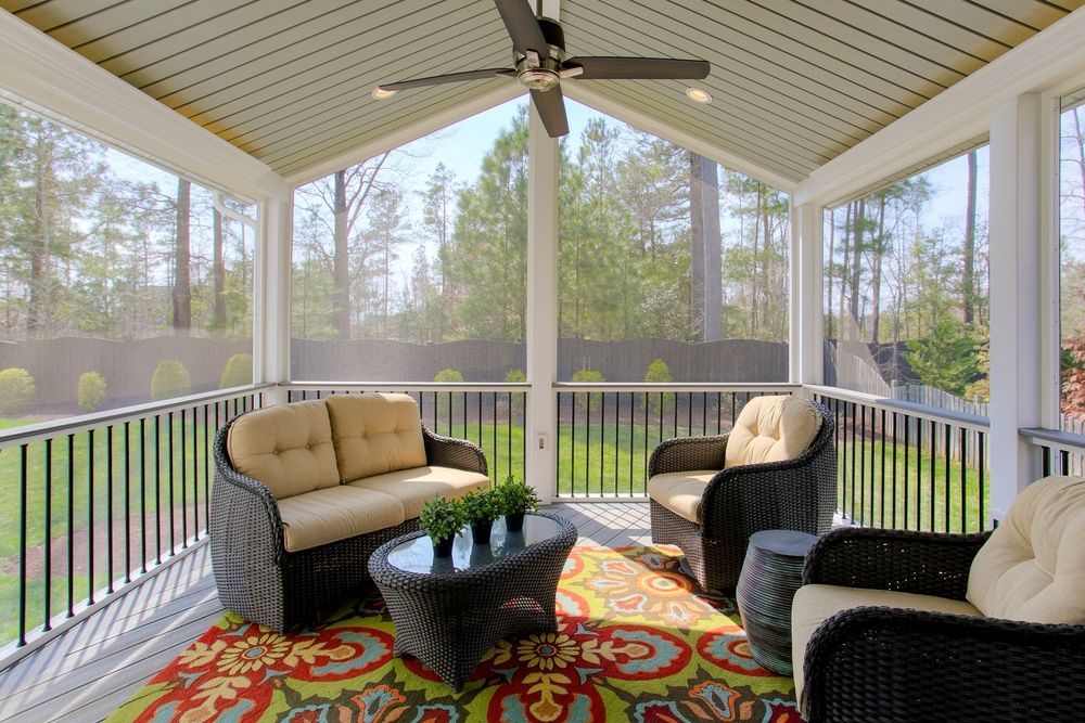 A furnished, screened-in porch with wicker seating, a patterned area rug, and a ceiling fan overlooking a wooded backyard.