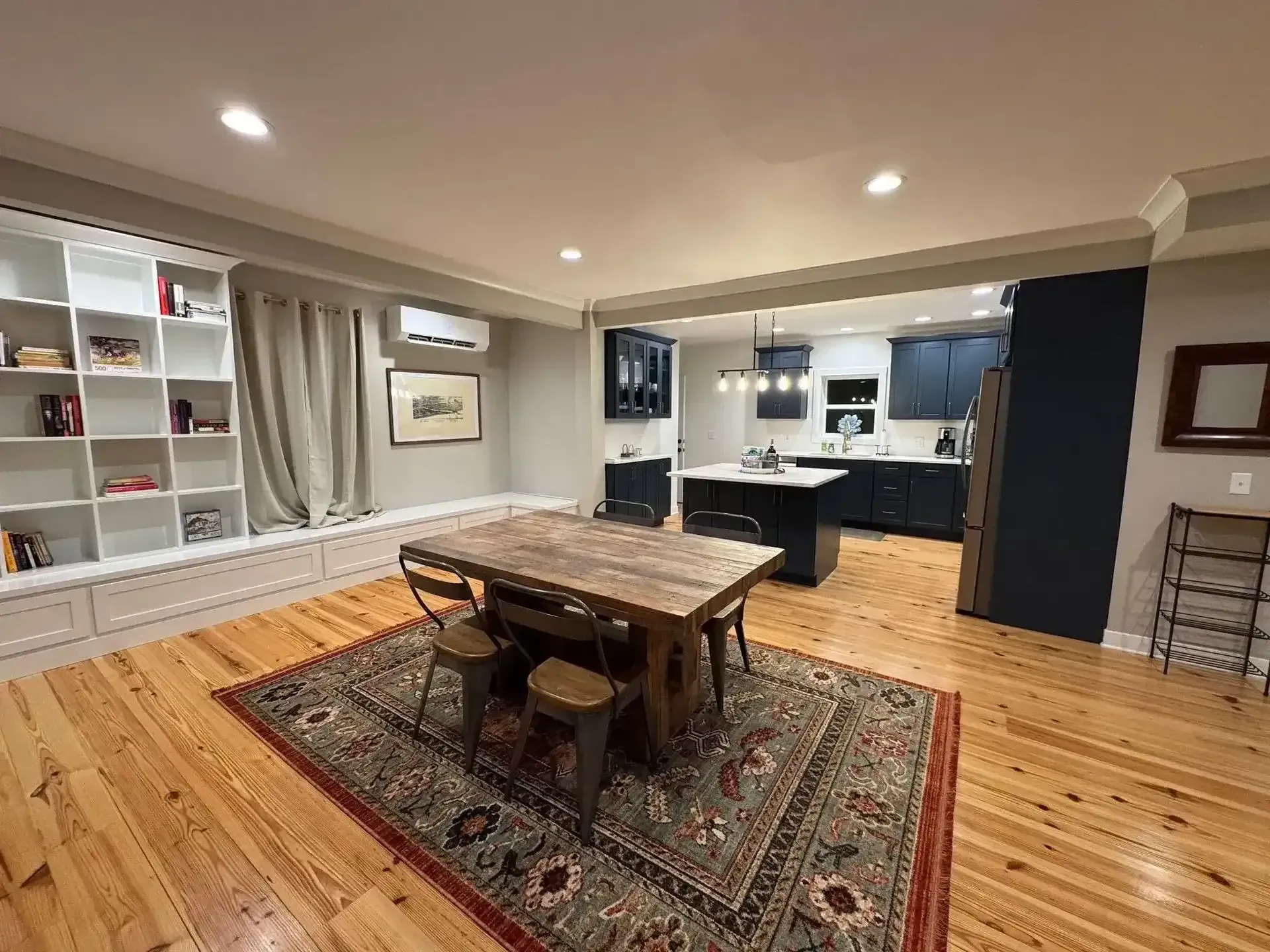 Open living room and kitchen with wood floors, dining table on rug, shelves, and recessed lighting.