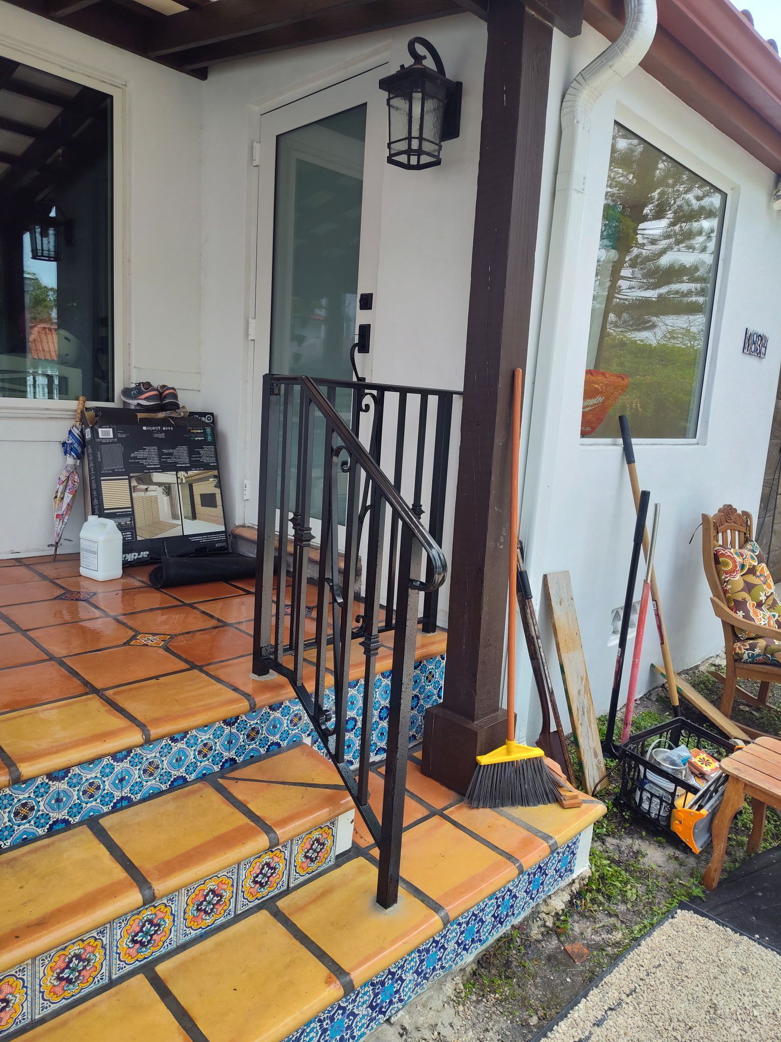 Porch with terracotta steps decorated with blue tiles, a black iron handrail, and various items stored near the door.