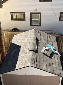 View from above of a house roof with shingles and a roof vent near a white exterior wall and windows.