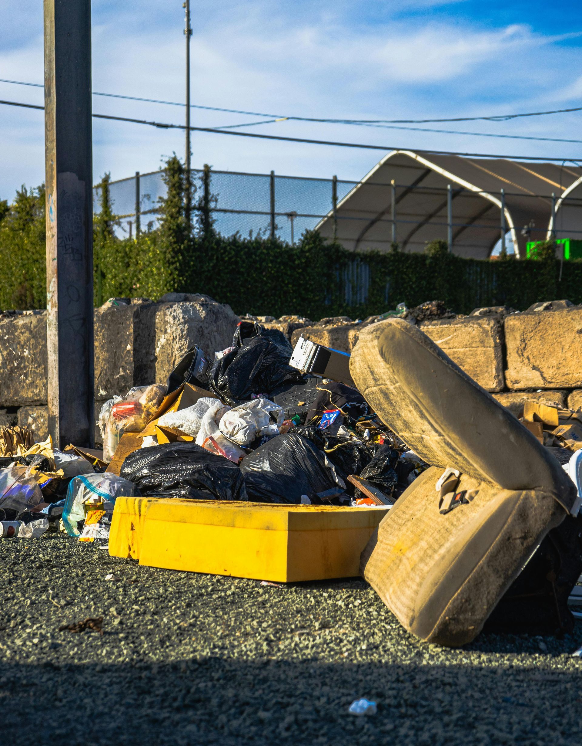 Overflowing trash bags and a broken yellow mattress on gravel beside a stone wall outdoors
