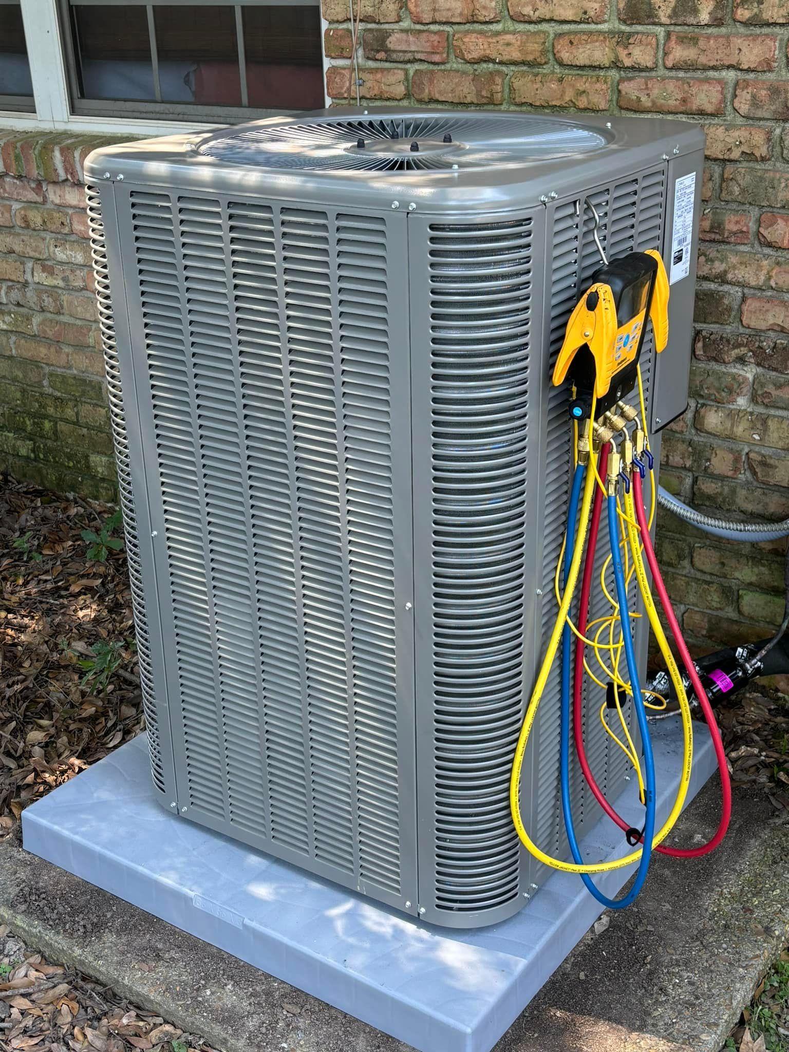 Air conditioning unit, gray metal, with gauges attached, sitting on a gray pad next to a brick wall.