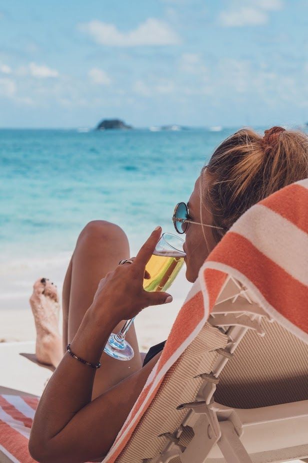 Woman relaxing on a beach, sipping a drink, ocean in the background. Orange and white striped towel.