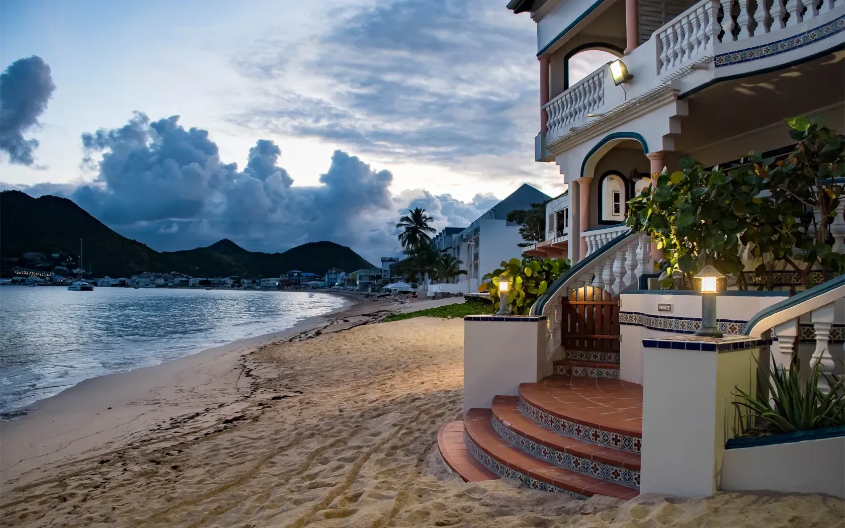 Beachfront villa at dusk with steps leading to the entrance and a view of mountains and the sea.