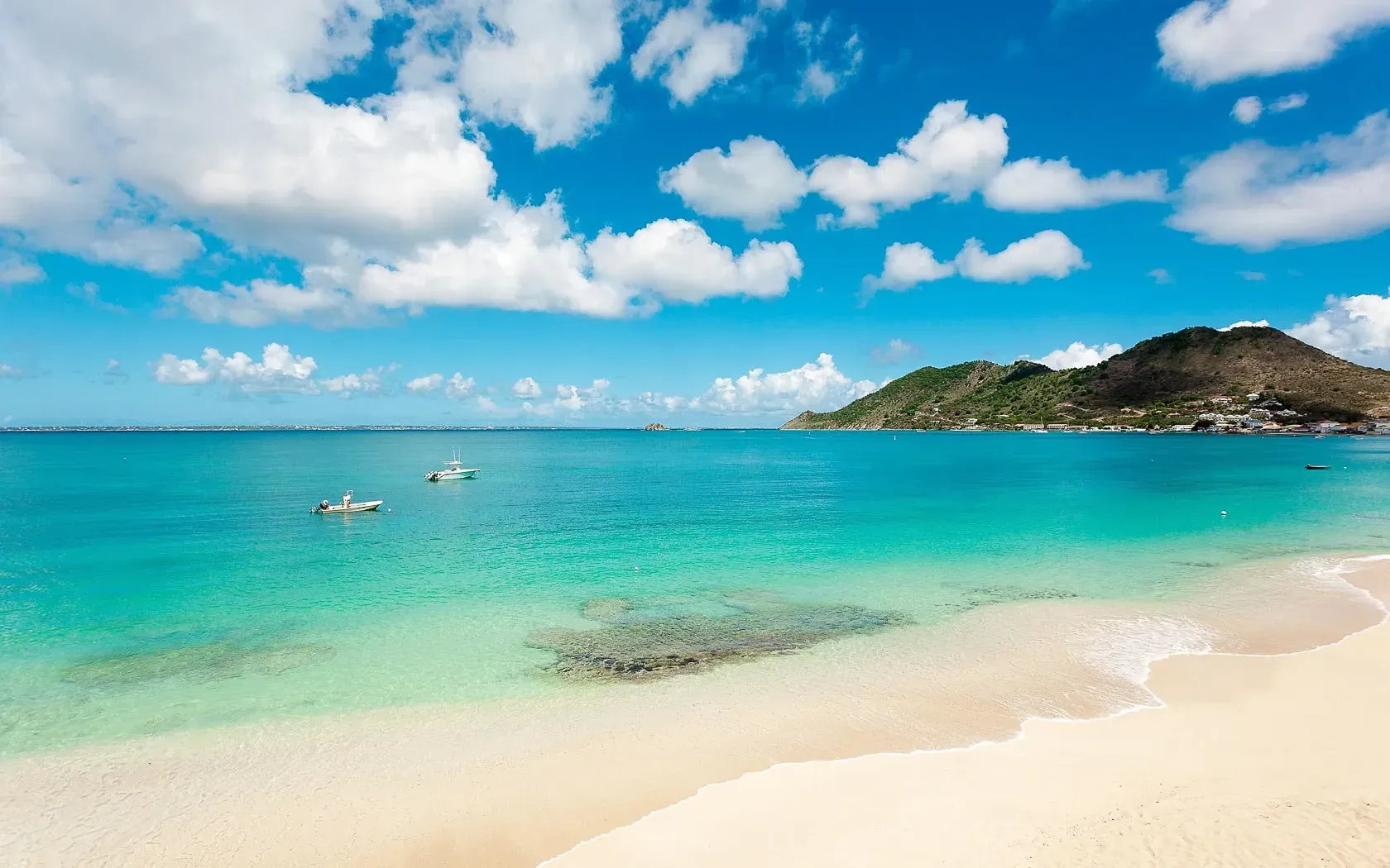Beach with turquoise water, white sand, blue sky, clouds, boats, and a green hill in the background.
