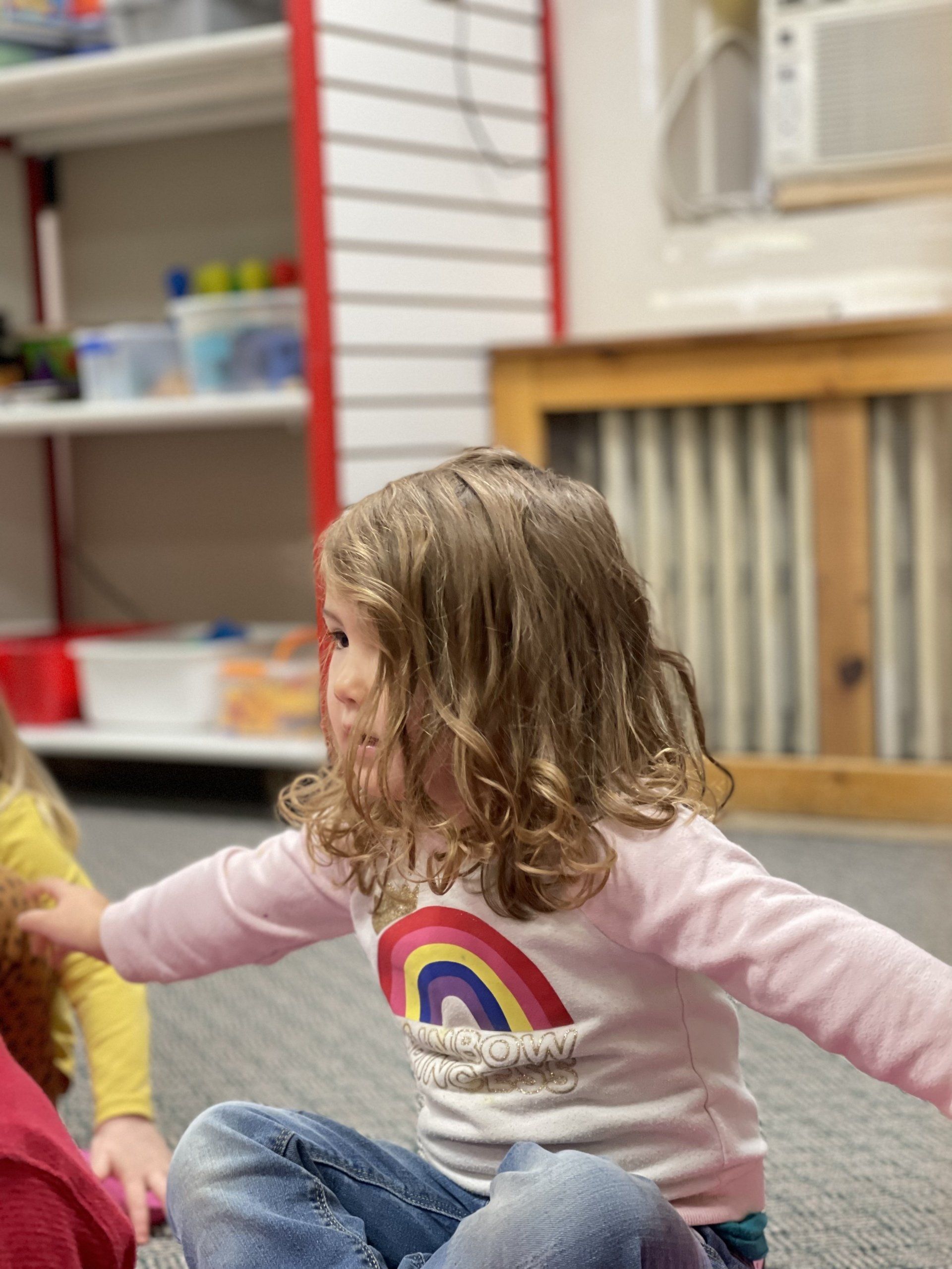 Young child with curly hair wearing a pink shirt with a rainbow graphic, sitting on the floor.