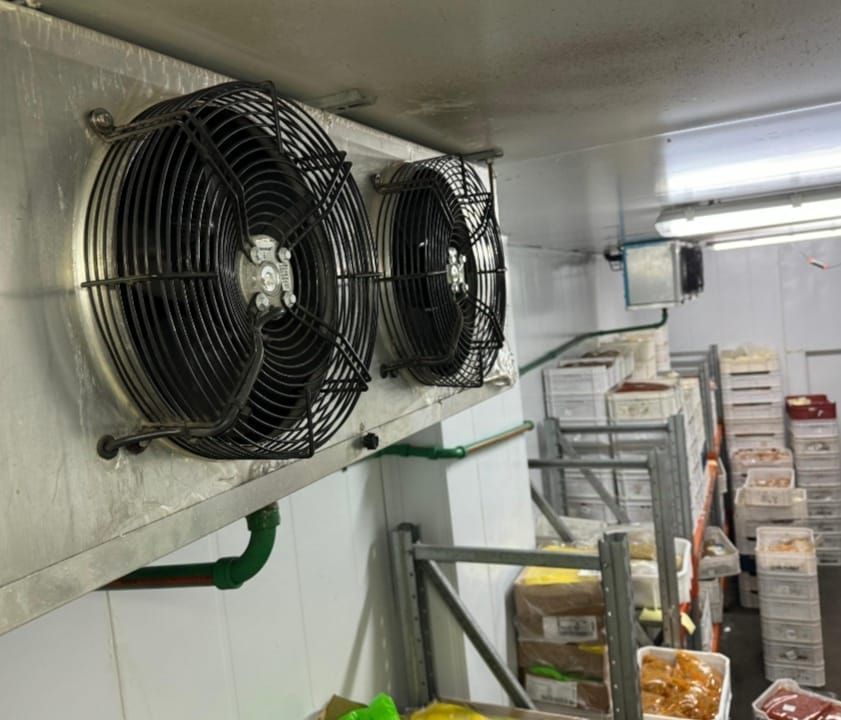 Dos ventiladores industriales montados en una pared en una cámara frigorífica, con cajas de mercancías visibles.