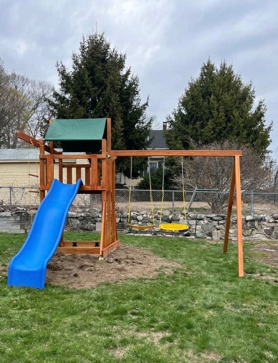 Wooden backyard playset with blue slide and swing frame on grass, with trees and stone wall behind.