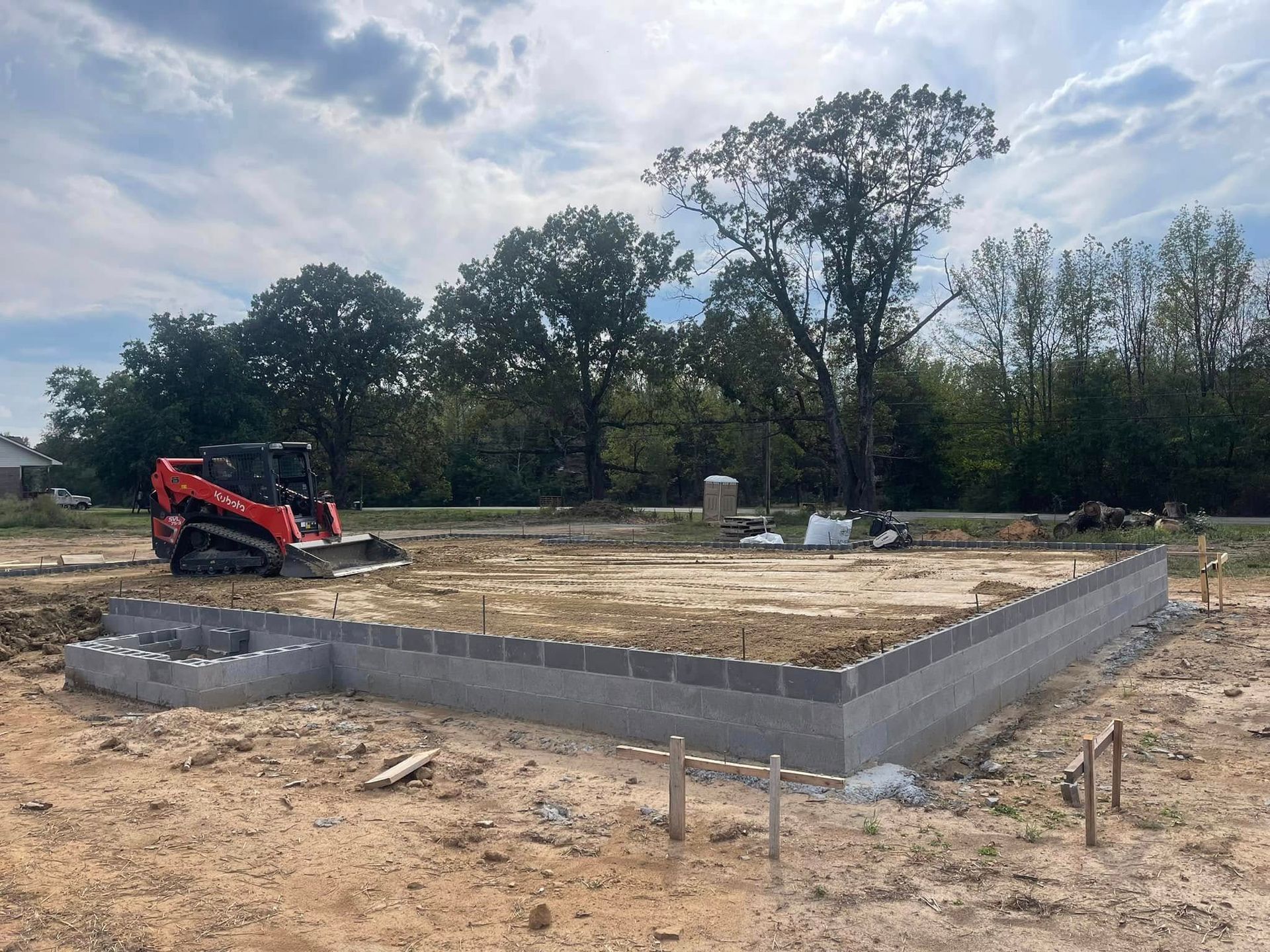 A red skid steer loader parked next to a concrete block foundation under construction on a dirt lot with trees behind it.