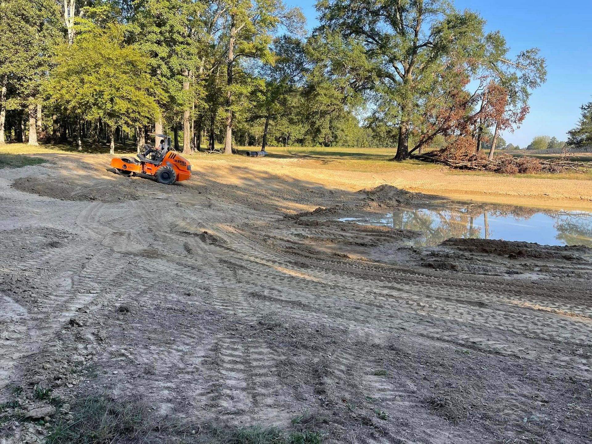 An orange piece of construction machinery operates on a dirt site near a small pond surrounded by trees.