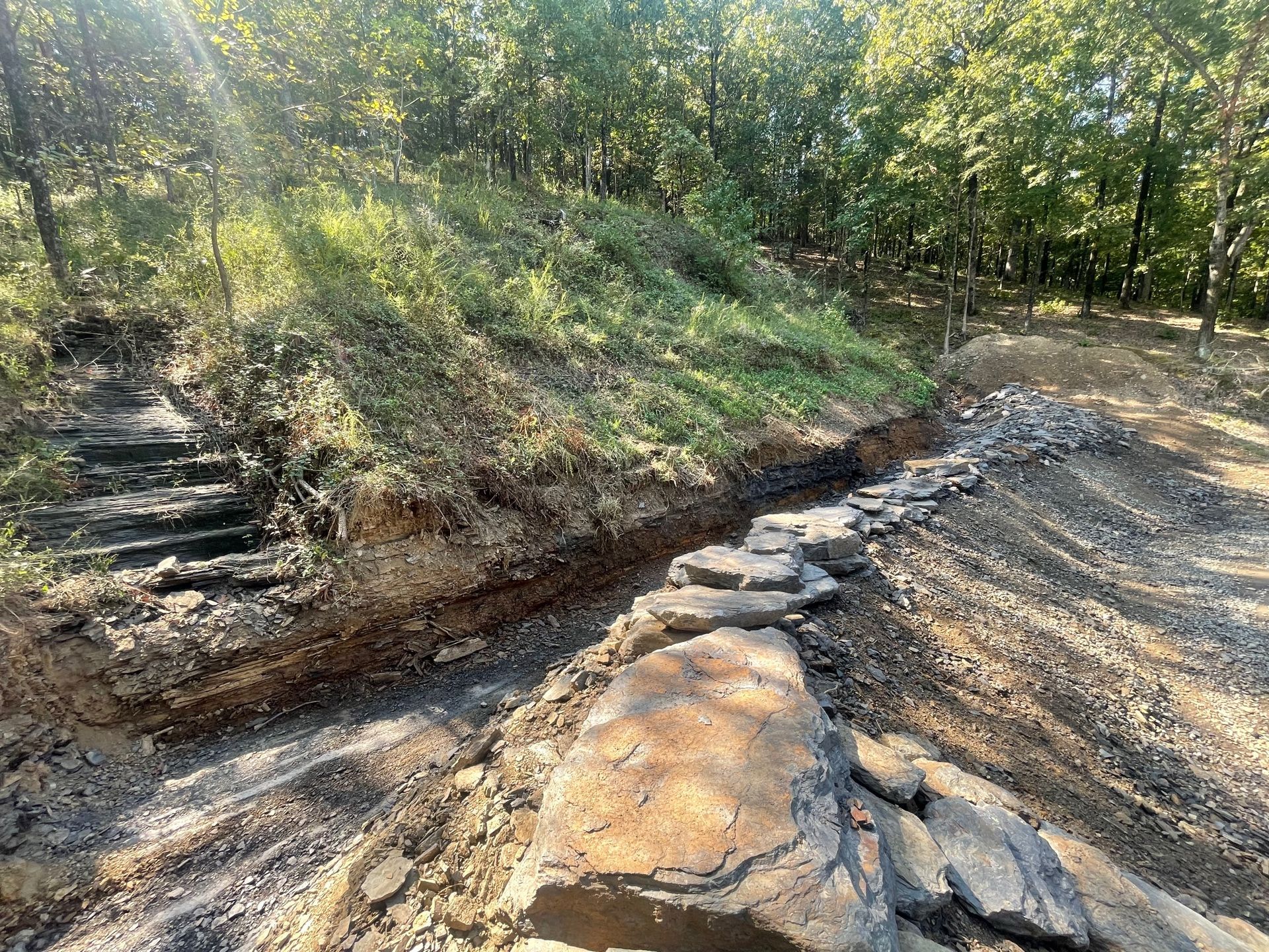 A partially constructed stone retaining wall along a slope at the edge of a wooded area.