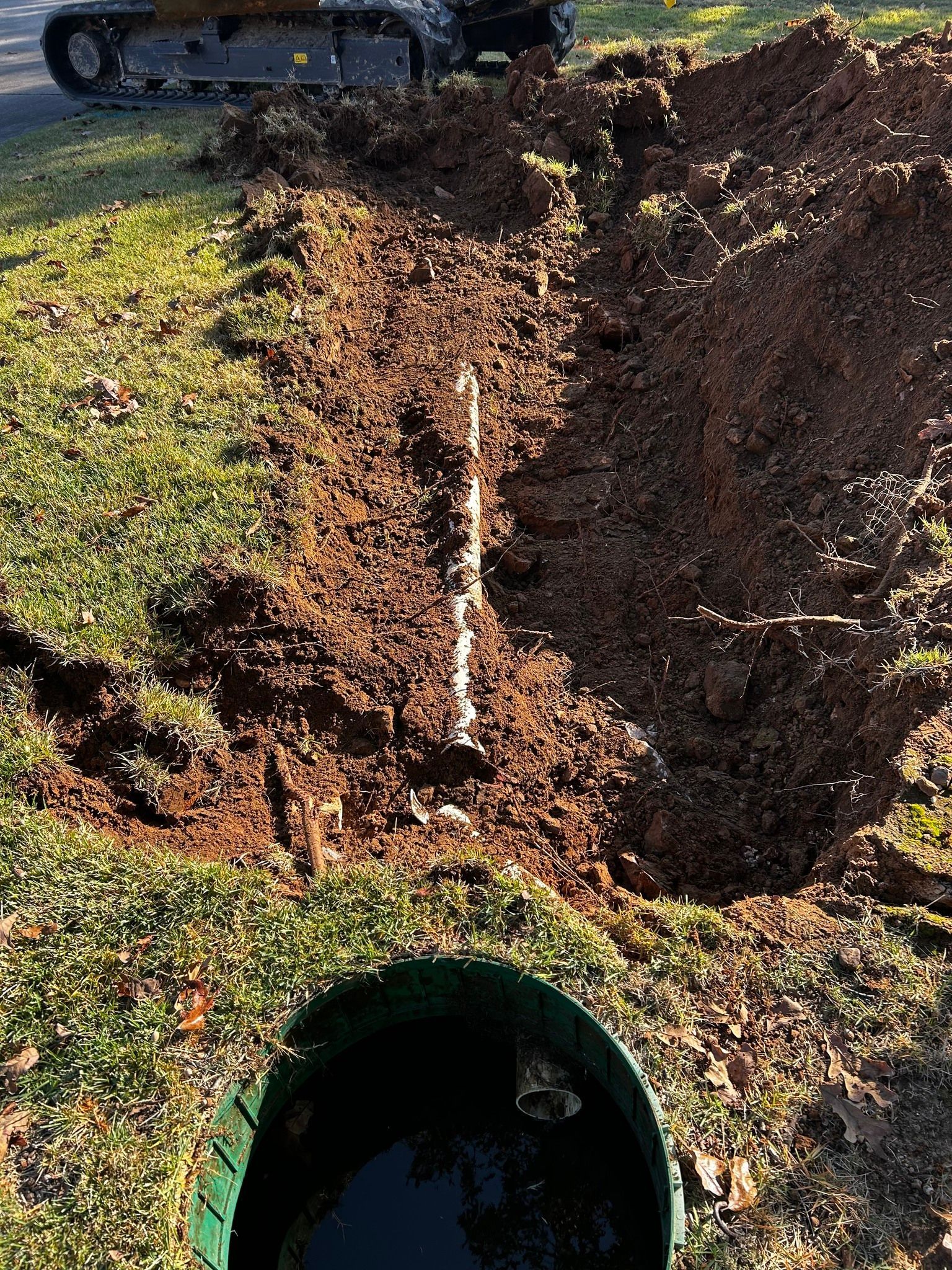 A track loader sits beside a freshly dug trench in a grassy yard, with a pipe leading to an open green drainage basin.