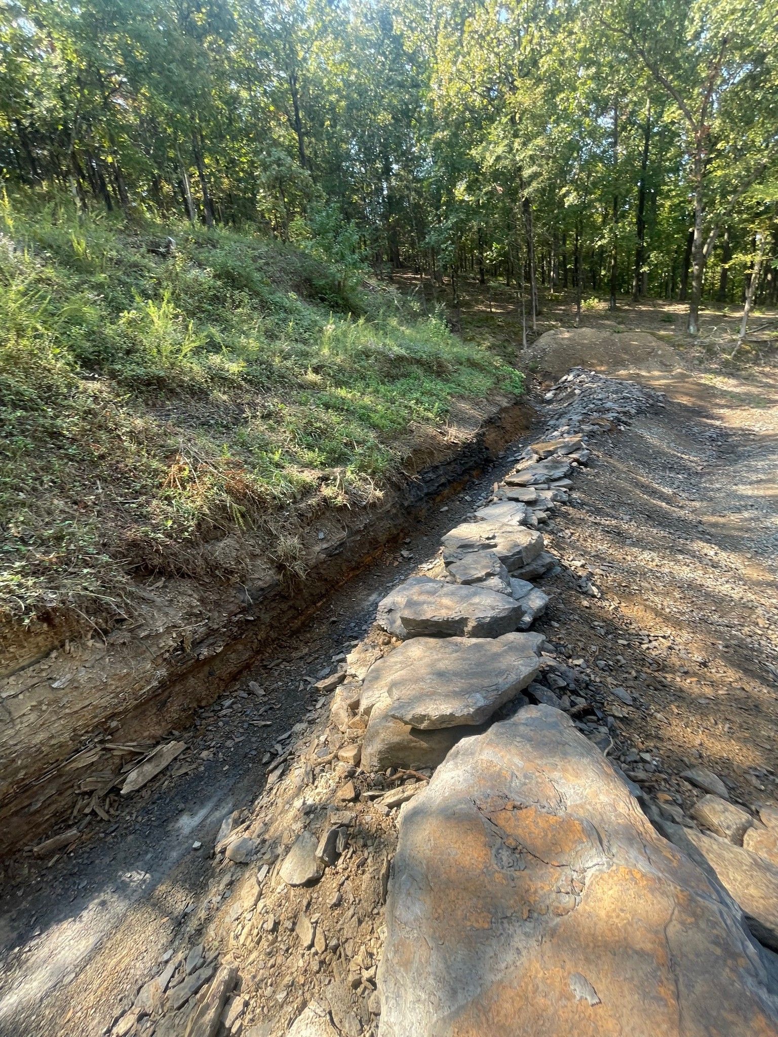 A line of large, flat boulders placed as a base for a retaining wall in a trench beside a wooded hillside.