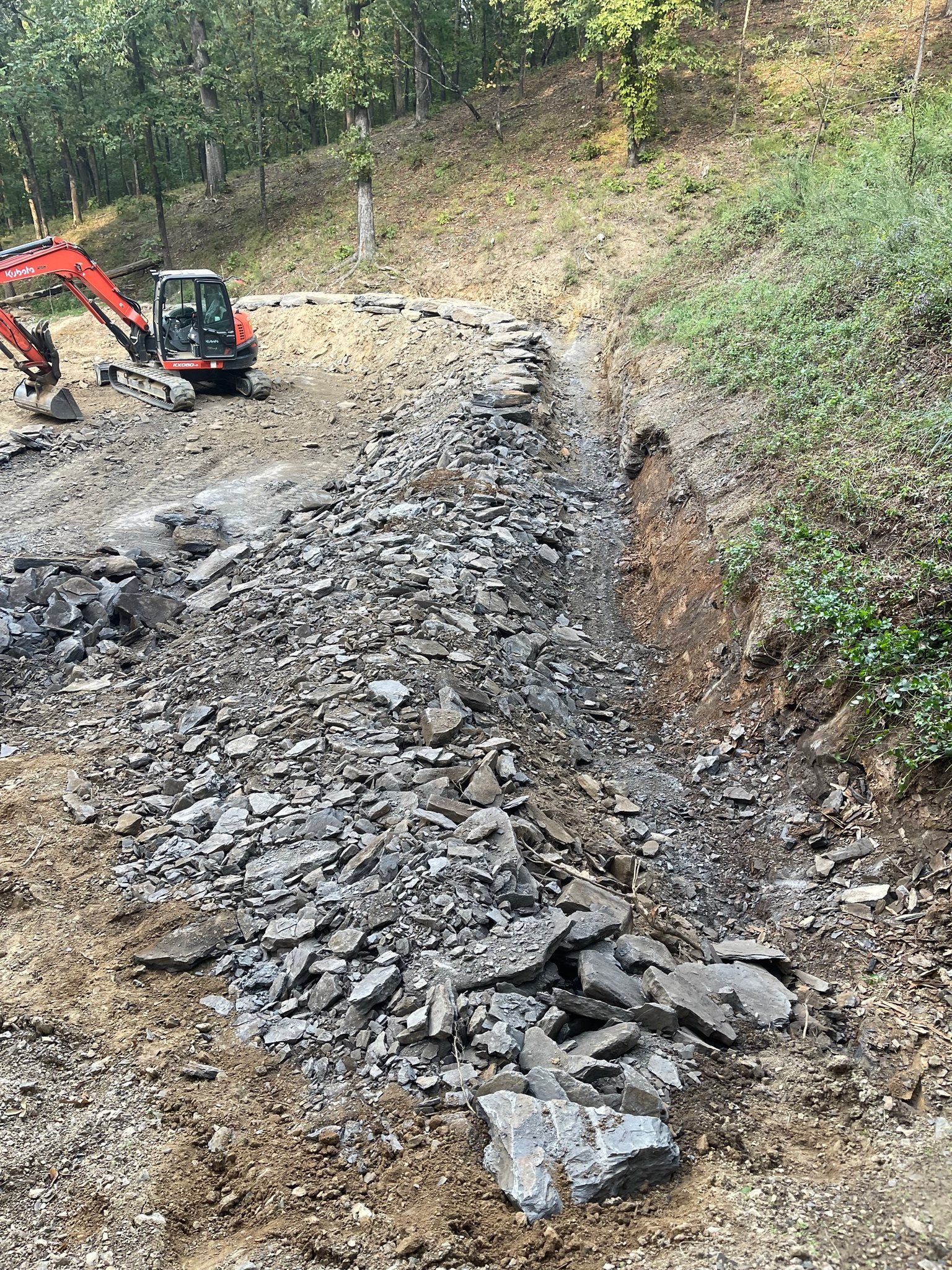 An orange excavator sits on a dirt hillside next to a large pile of grey crushed rock and stone.