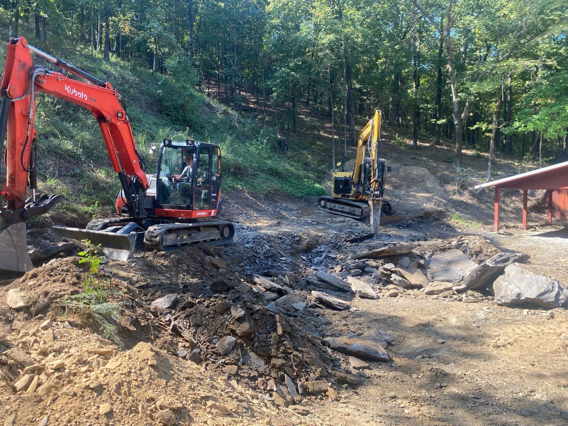 Two excavators, one orange and one yellow, work on clearing rocky terrain near a wooded hillside and a small building.
