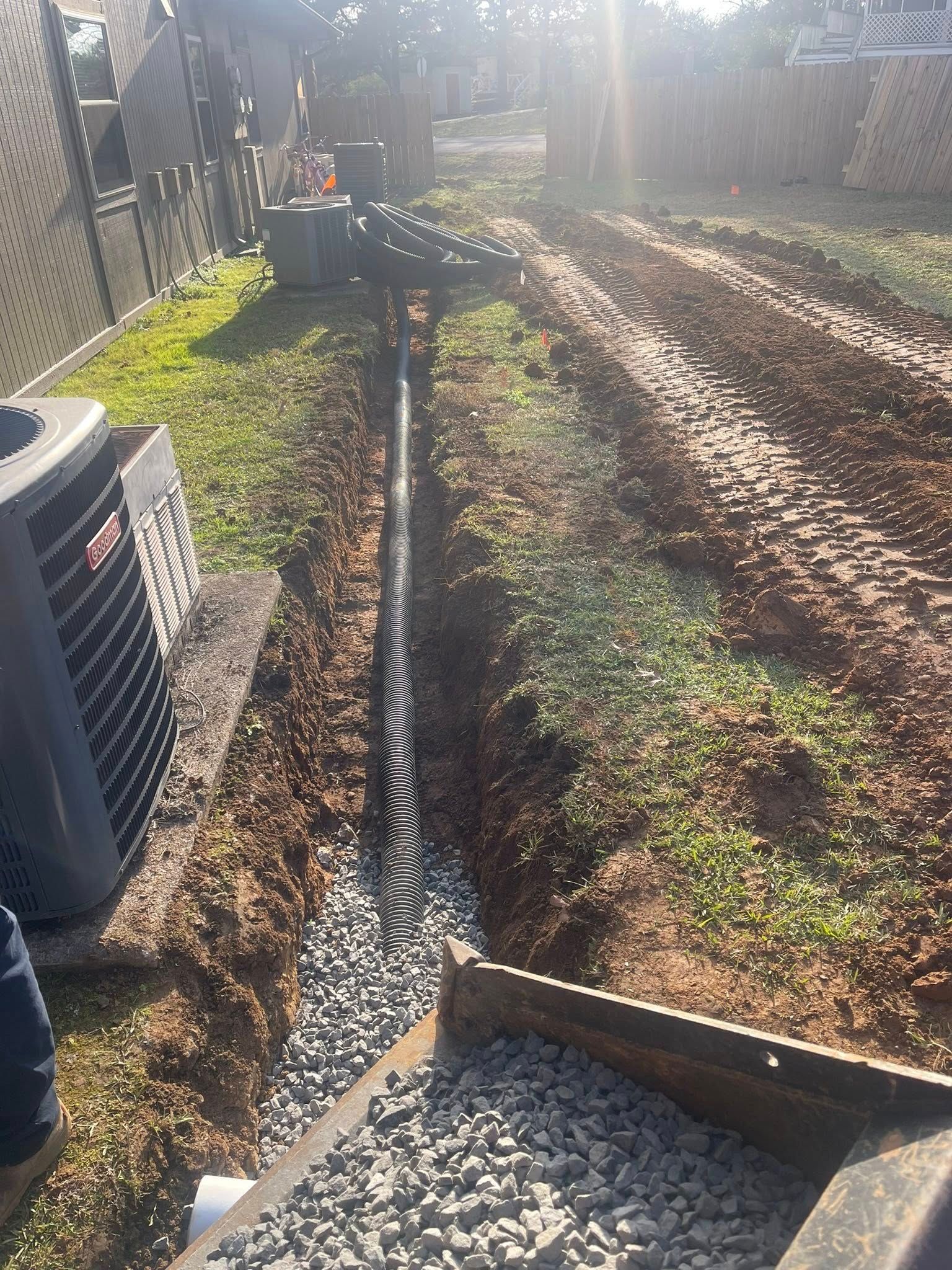 A black drainage pipe sits in a dirt trench filled with gravel, next to an outdoor air conditioning unit.