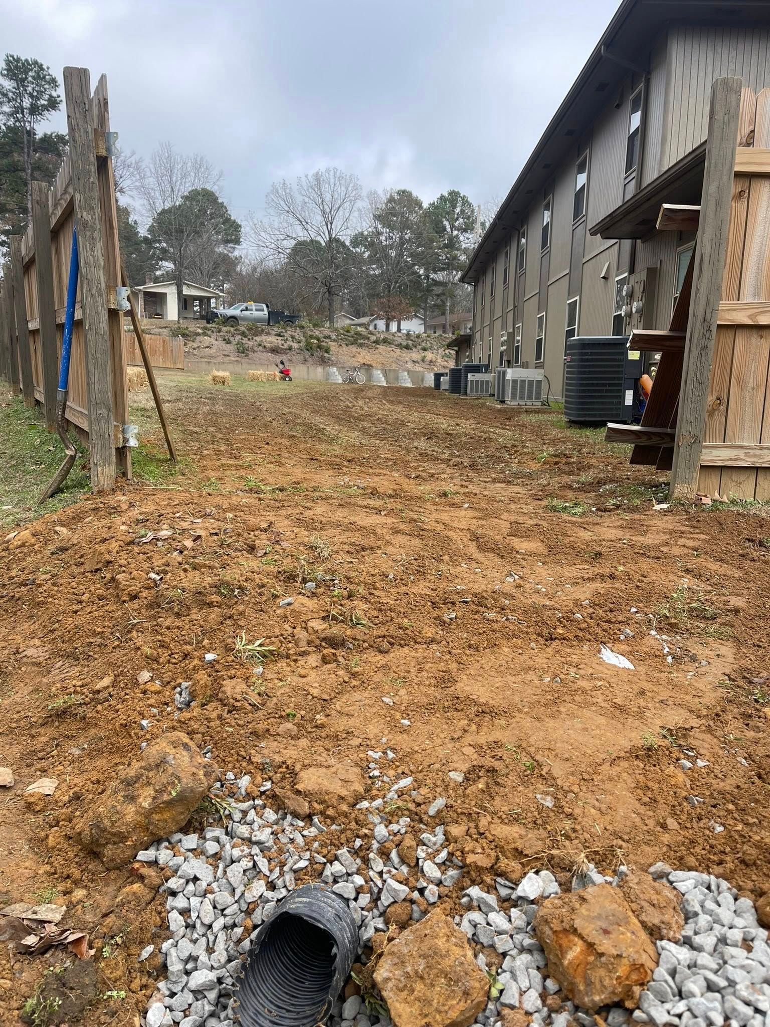 A dirt-filled construction area between a wooden fence and a building, featuring a black drainage pipe and gravel.