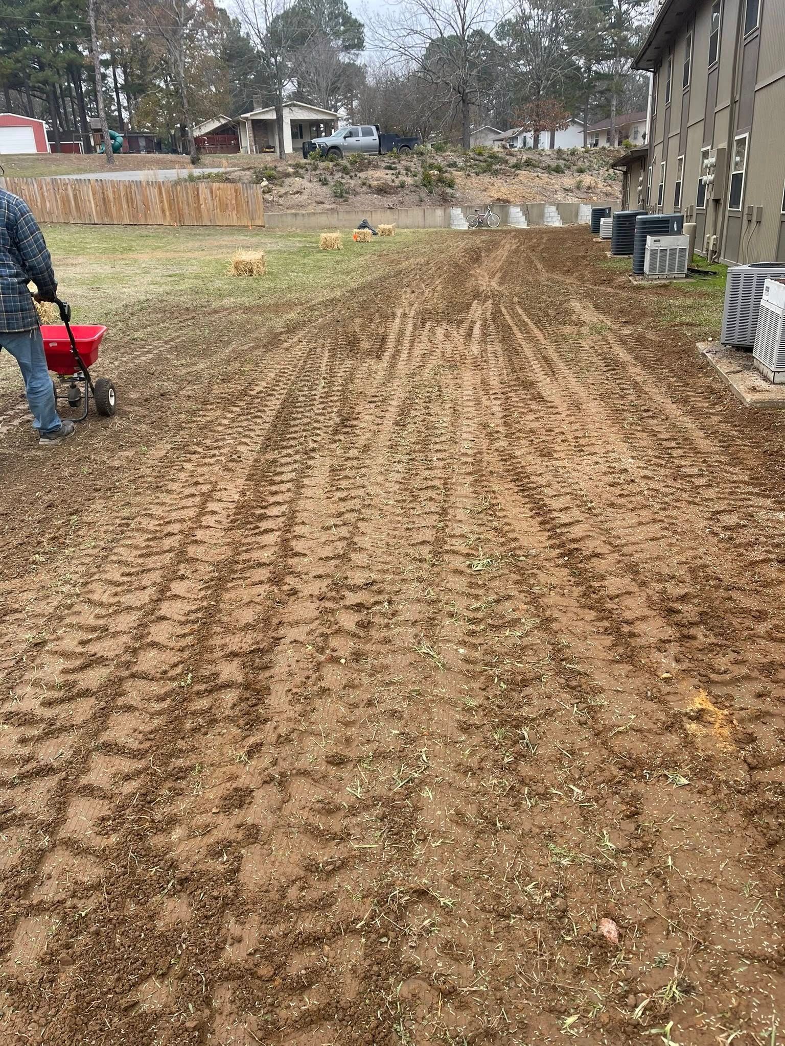 A person uses a red walk-behind fertilizer spreader on a muddy, dirt-covered yard alongside an apartment building.