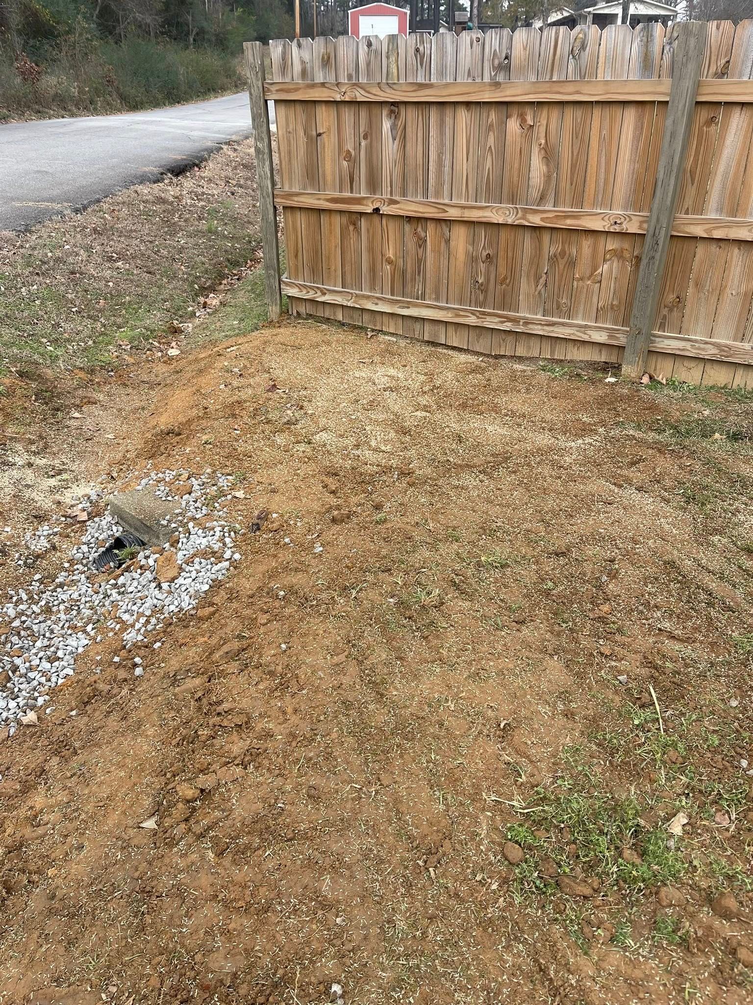 A wooden privacy fence borders a dirt and gravel yard with a small drainage area featuring light-colored stones.