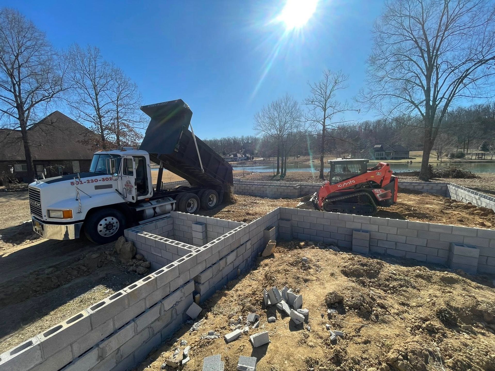 A white dump truck unloads dirt at a construction site with concrete block foundations and an orange skid-steer loader.