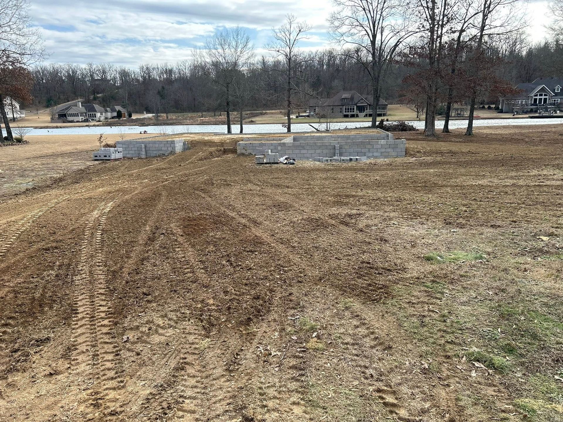 A construction site with concrete foundation blocks on a dirt lot facing a lake, trees, and houses in the distance.