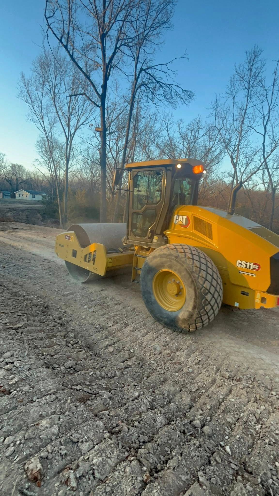 A yellow road roller sits on a dirt path in a wooded area under a clear blue sky.