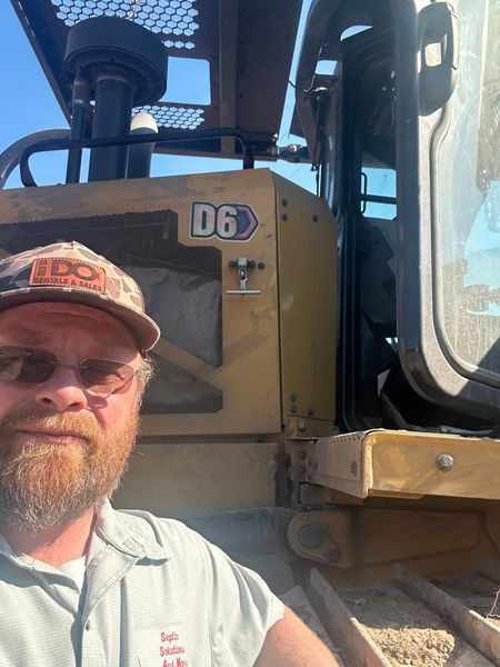 A man with a beard and baseball cap posing for a selfie in front of a yellow Caterpillar D6 bulldozer.