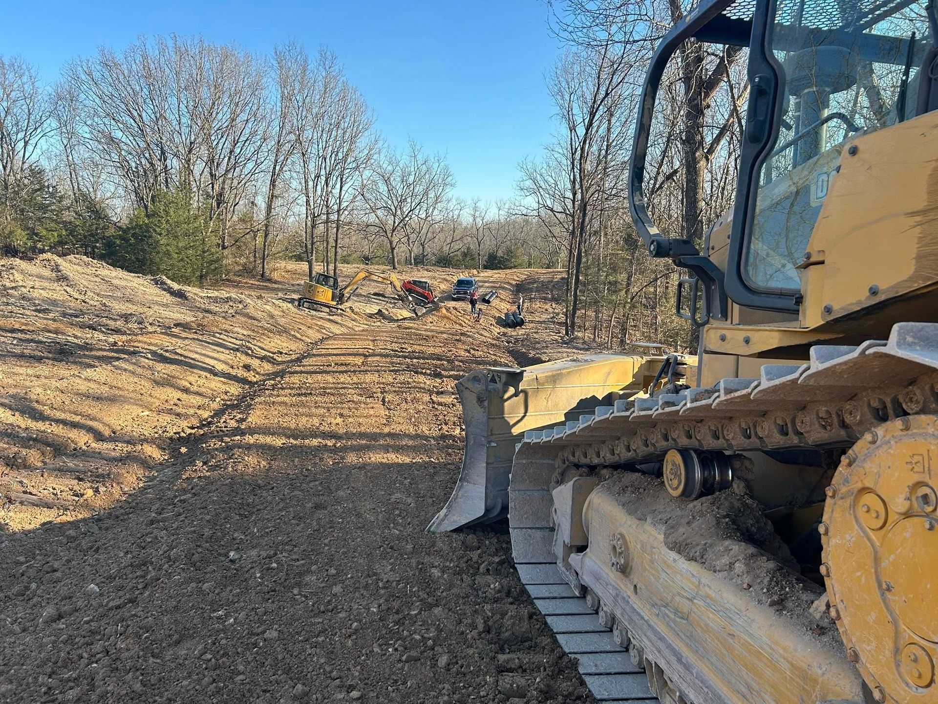 A yellow bulldozer on a dirt path in a wooded area under a clear blue sky.