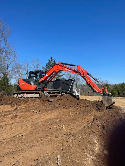 An orange excavator sits on a dirt mound in a field under a clear blue sky, working on a construction site.