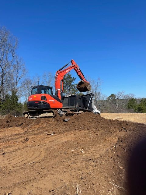 An orange excavator loads dirt into a dump truck on a dirt construction site under a clear blue sky.