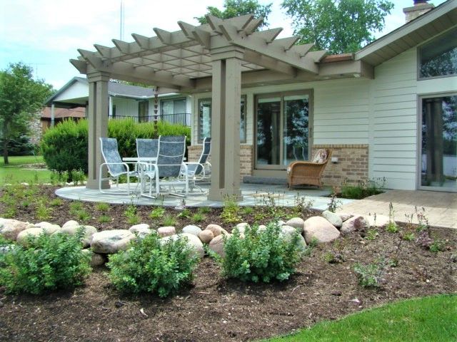 Patio with white pergola, outdoor table and chairs beside a house, surrounded by landscaped garden beds