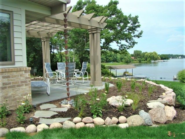 Patio with white chairs and pergola beside a landscaped lakefront garden and calm water