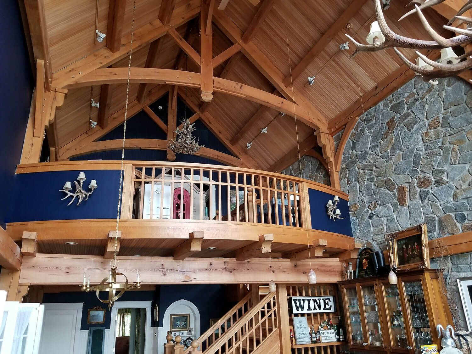 Rustic lodge interior with timber beams, stone wall, balcony, stairs, and warm wood decor