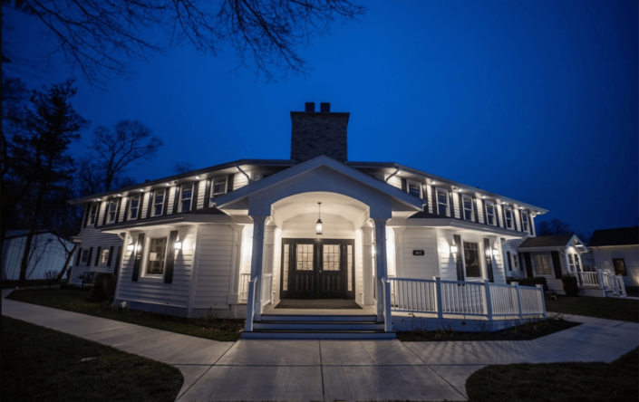 White house with wraparound porch lit at dusk under a deep blue sky