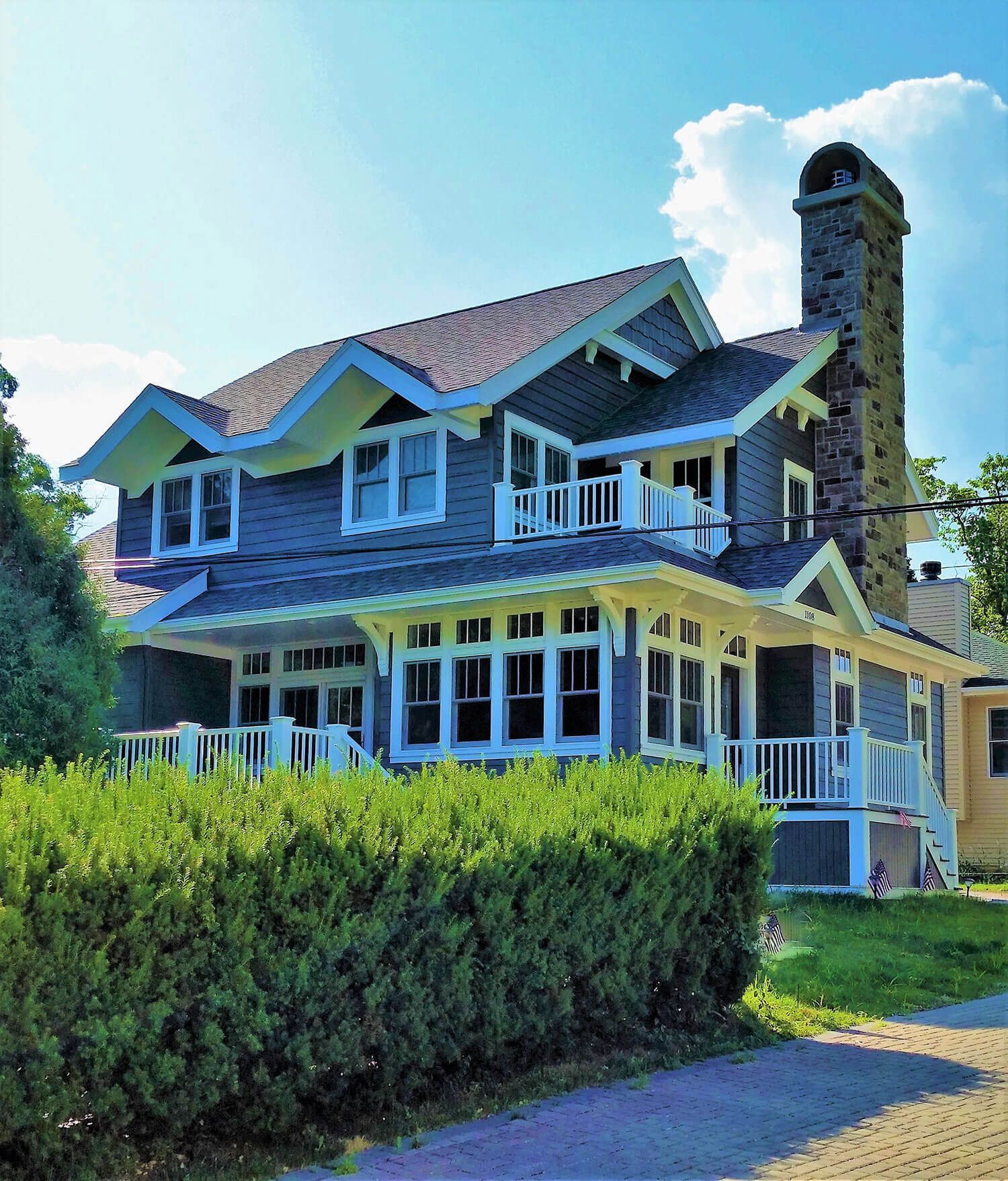 Large blue-gray house with white trim, wraparound porch, tall chimney, and green hedges under a sunny sky