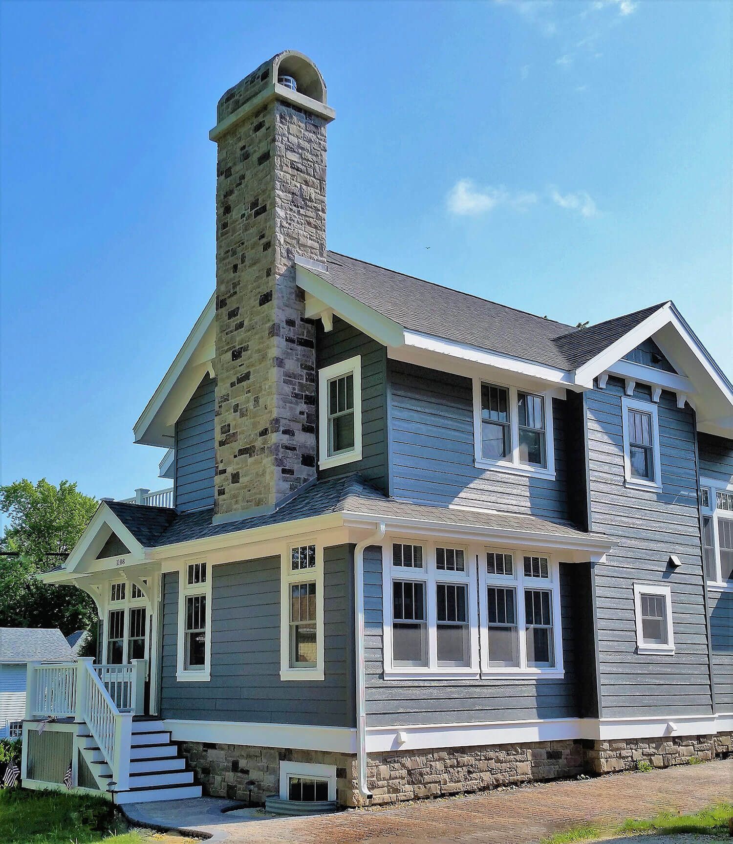 Blue-gray Victorian-style house with stone chimney, white trim, and wraparound porch under a clear sky