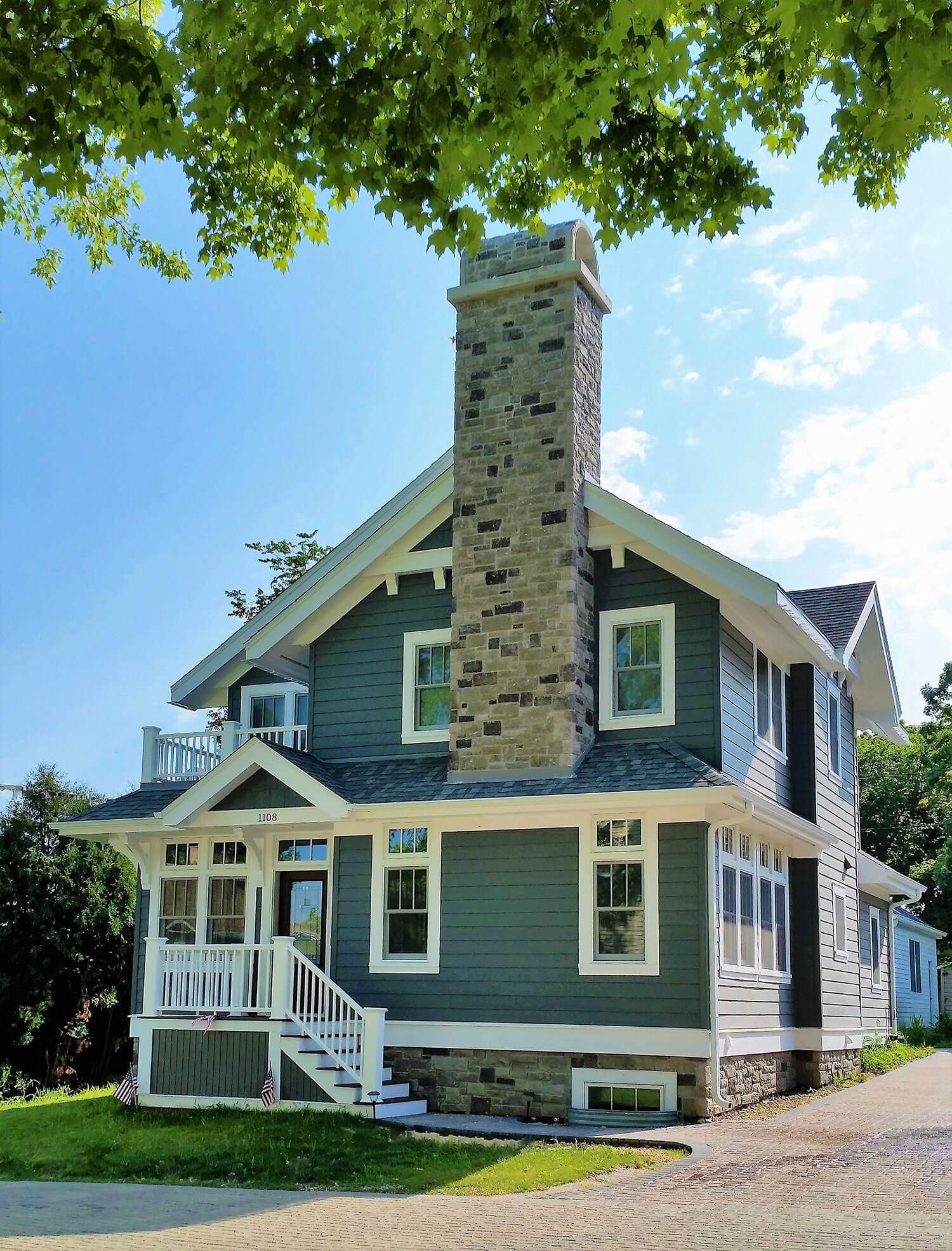 Blue house with stone chimney and white trim under leafy tree branches on a sunny day