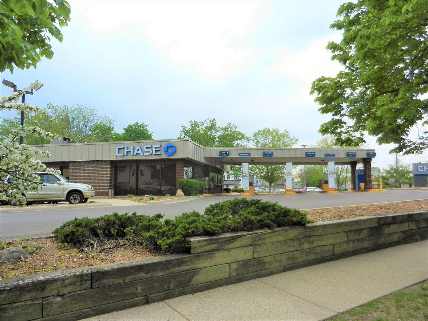 Chase bank branch with drive-thru lanes beside a sidewalk and stone planter under leafy trees