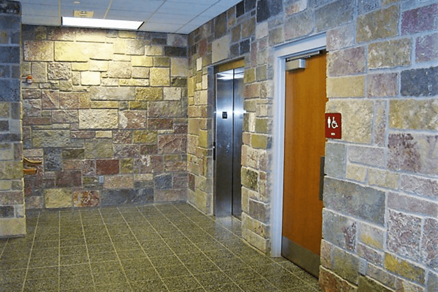 Stone-walled hallway with tiled floor and two wooden doors, one marked red, under bright ceiling lights