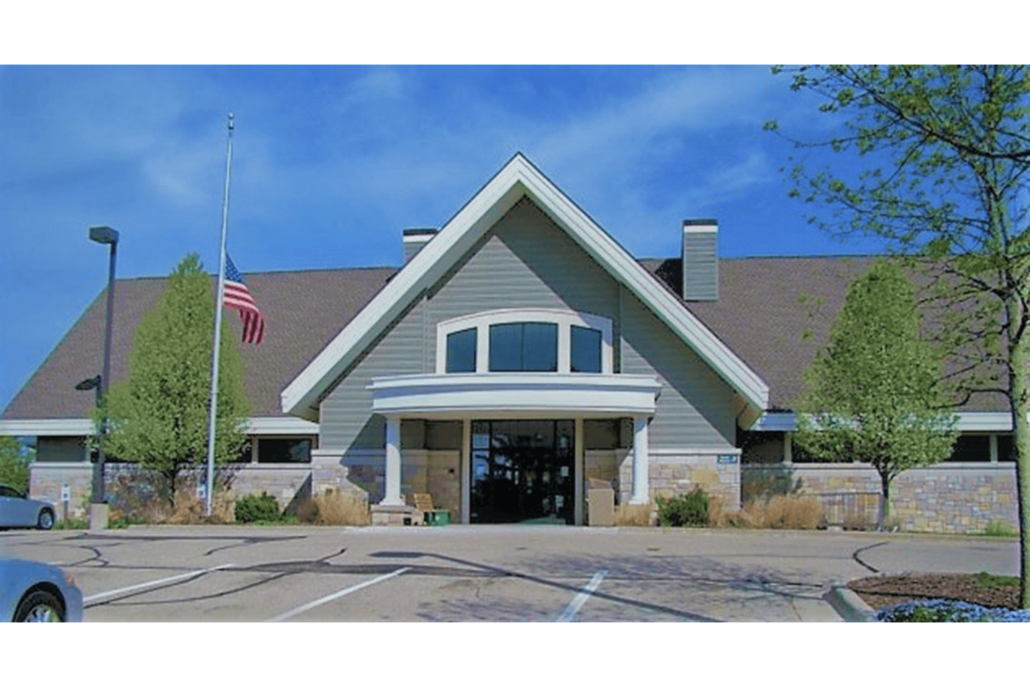 Front view of a beige community building with a peaked roof, glass entrance, and an American flag by the parking lot