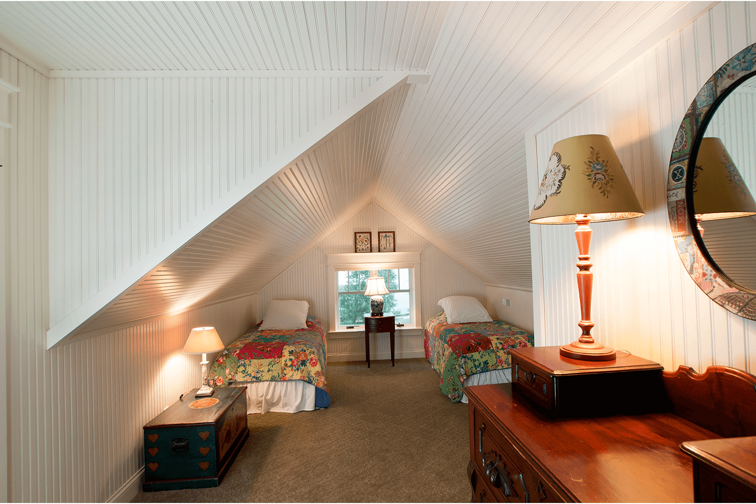 Small attic bedroom with two twin beds, sloped white walls, warm lamps, and a dresser with a mirror