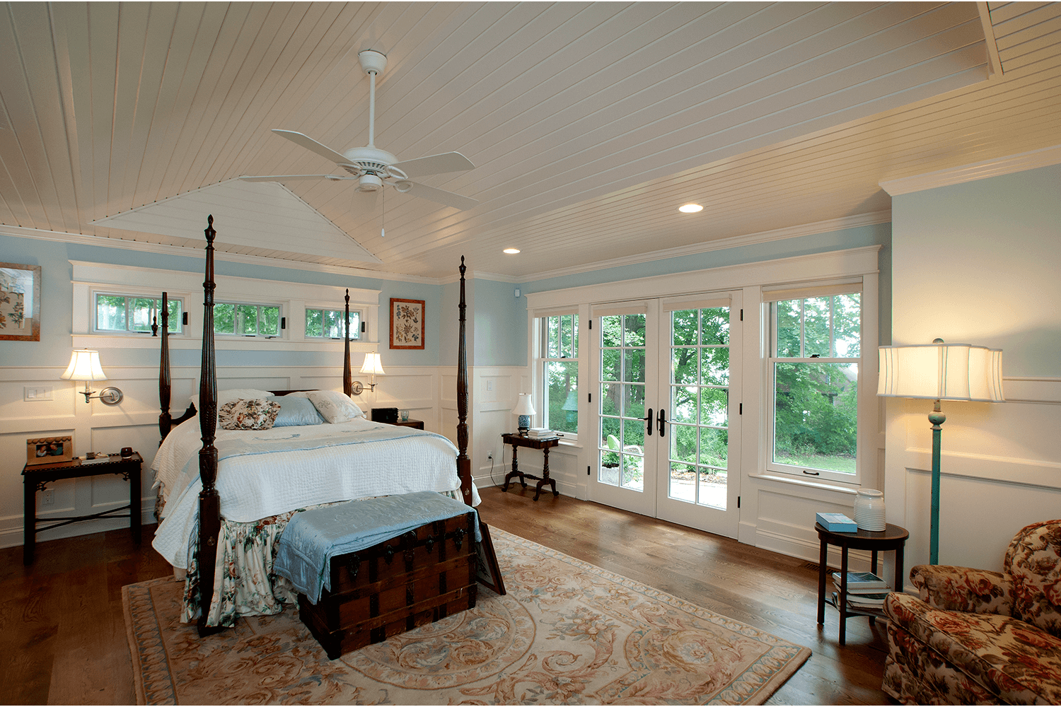 Bright bedroom with a large bed, wood floors, white ceiling fan, and windows overlooking greenery.